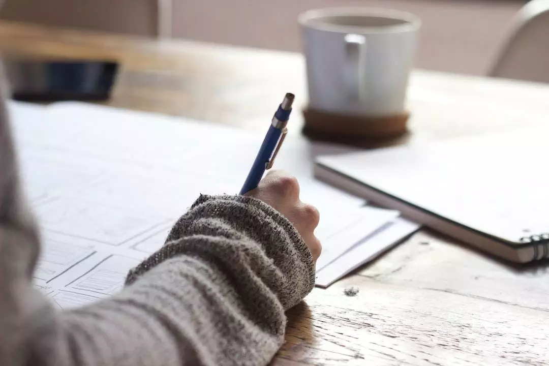 Person writing notes beside coffee mug on desk.